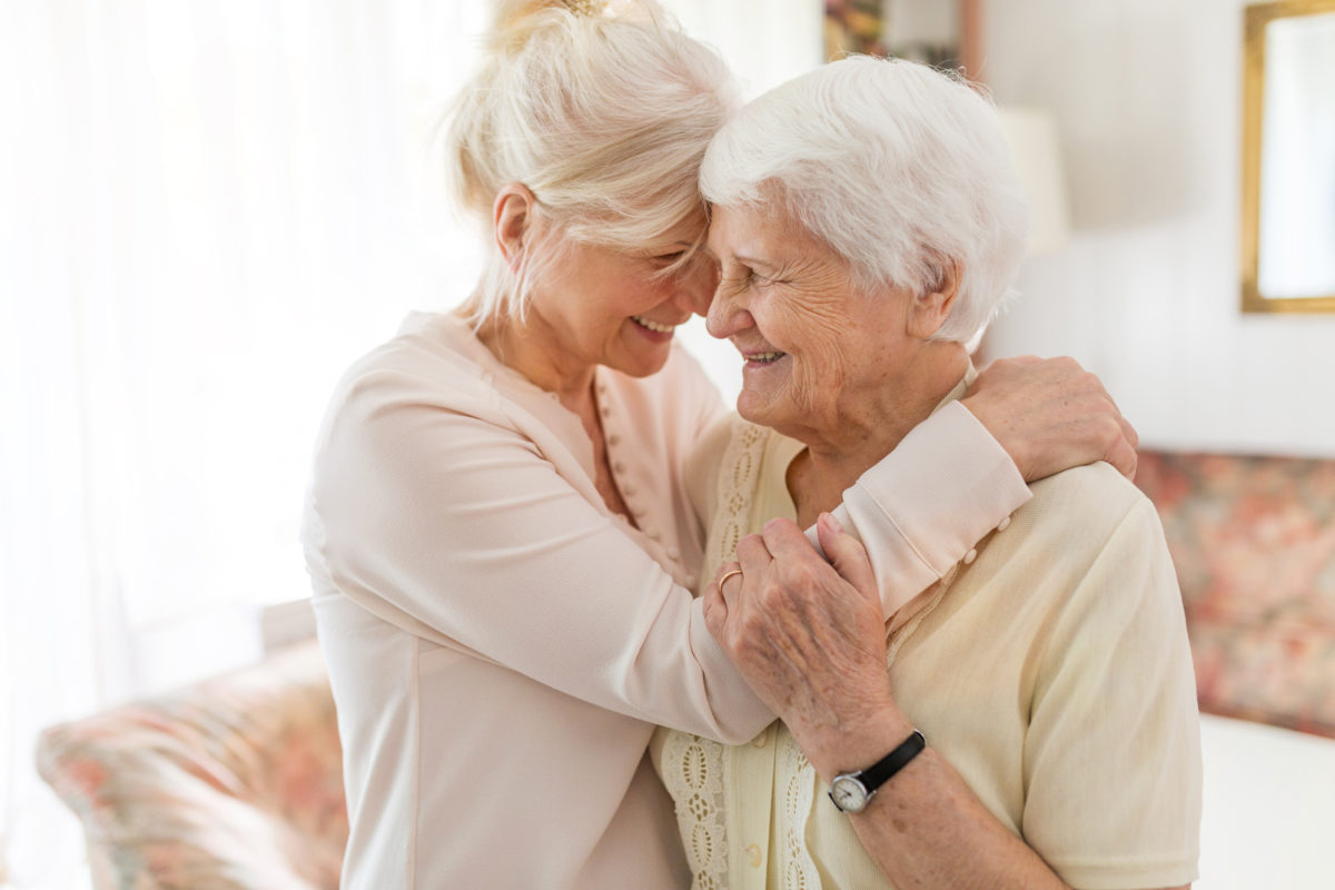 A smiling senior woman and her smiling daughter with their foreheads together. The daughter has her hand on her mother's shoulder, the mother has her hand on the daughter's arm.